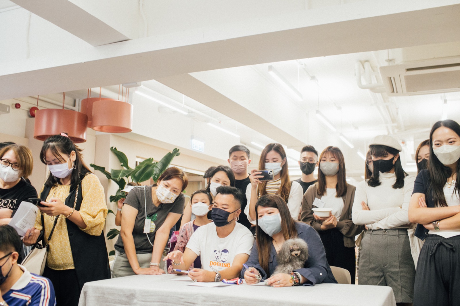 眾人圍在桌旁書寫，旁邊有人抱著小狗